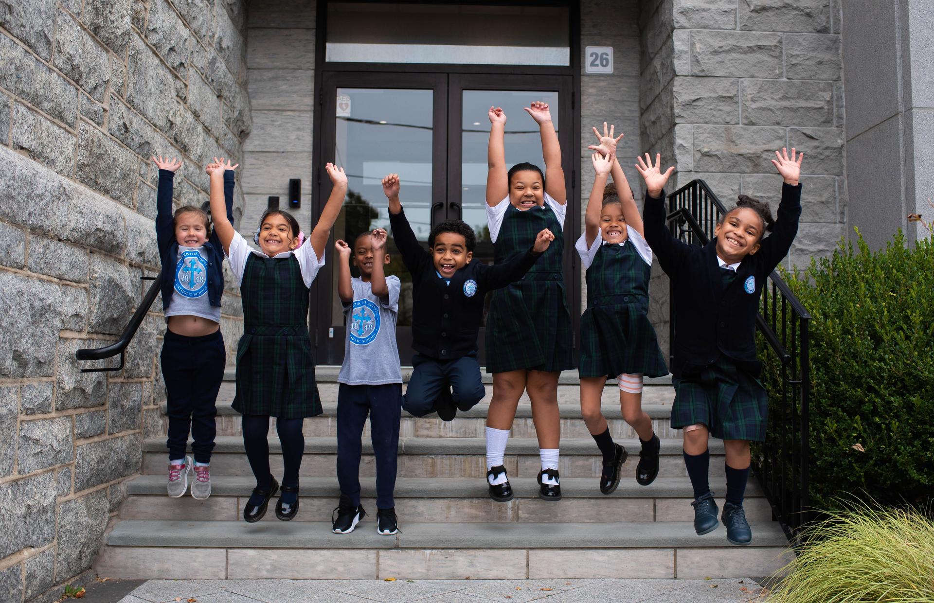 A group of children joyfully jumps on steps outside a building, expressing excitement.