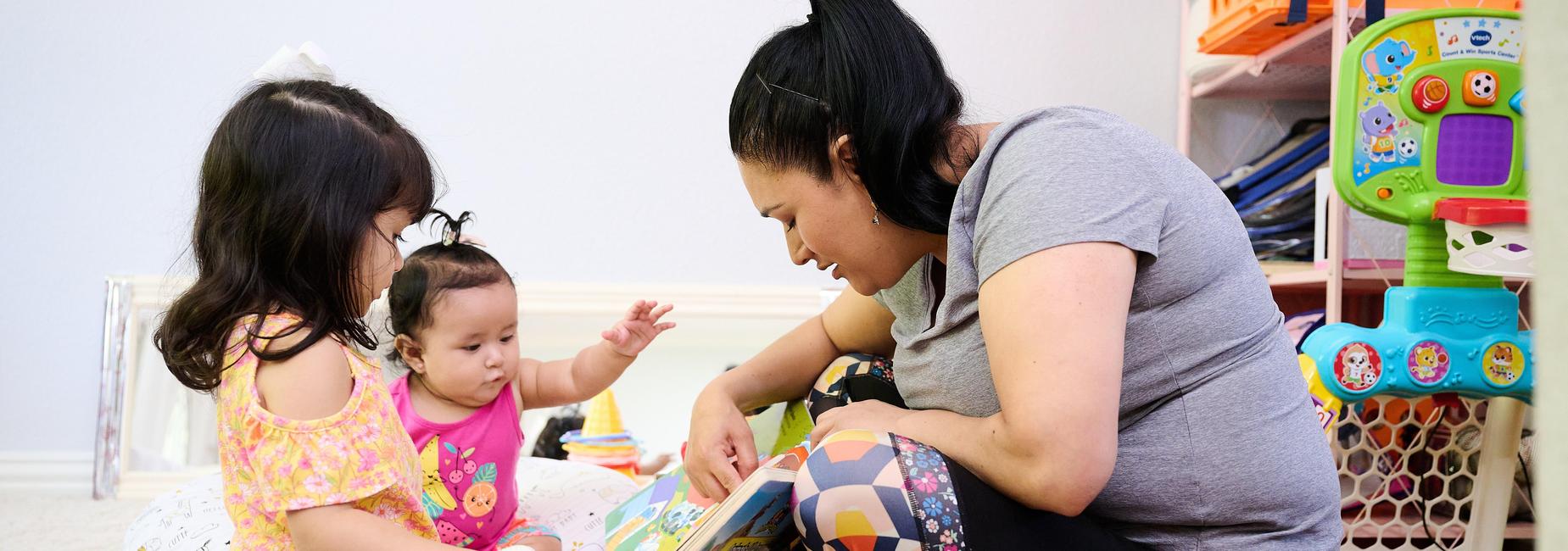 Woman reading a book to two young girls in a playroom setting.