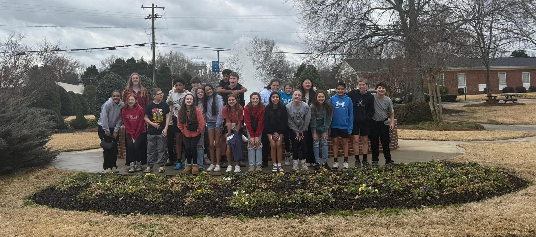 Group of students posing together outdoors near flower beds on a cloudy day.