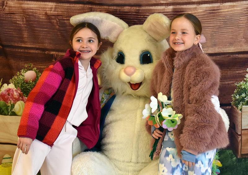 Two girls posing with a large bunny and flowers in a festive setting.