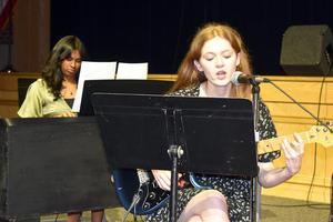 Young woman singing and playing guitar while another girl plays piano.
