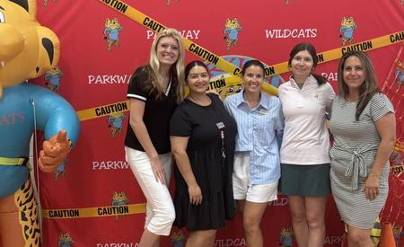 Six people pose together in front of a red wall with caution tape and a large cat mascot.