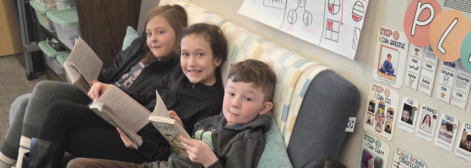Two children sit on a couch; one reads while another smiles at the camera.
