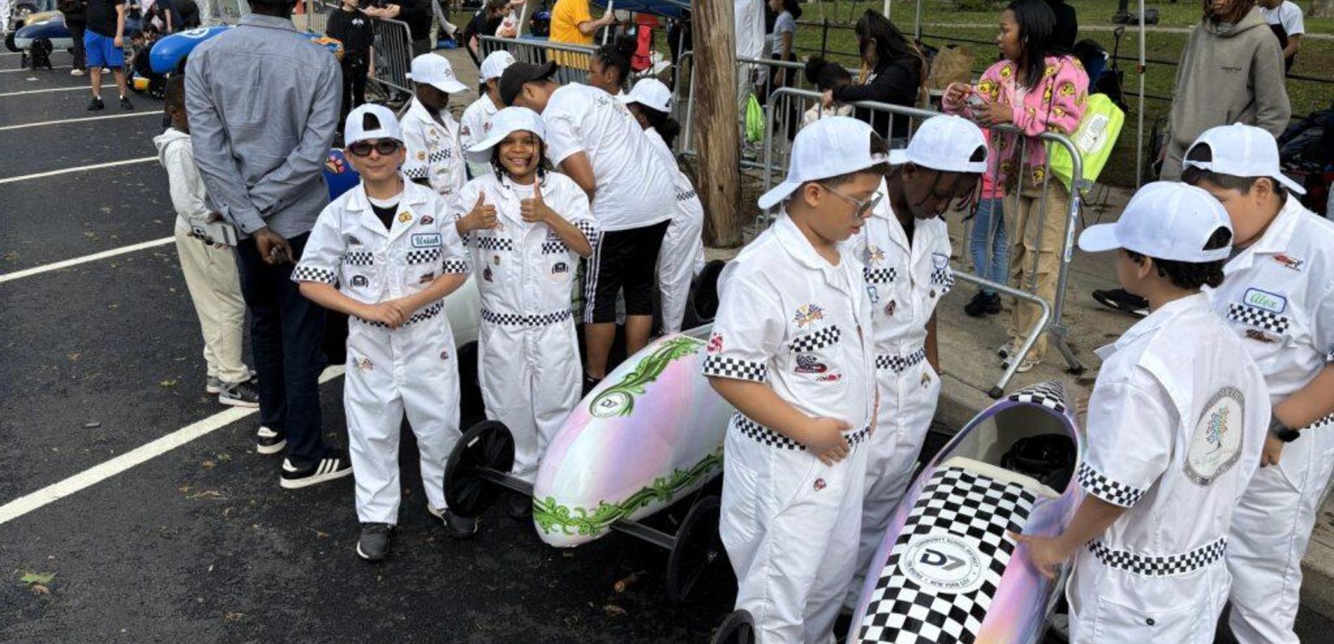 Children in racing suits smiling and posing next to soapbox cars at an event.