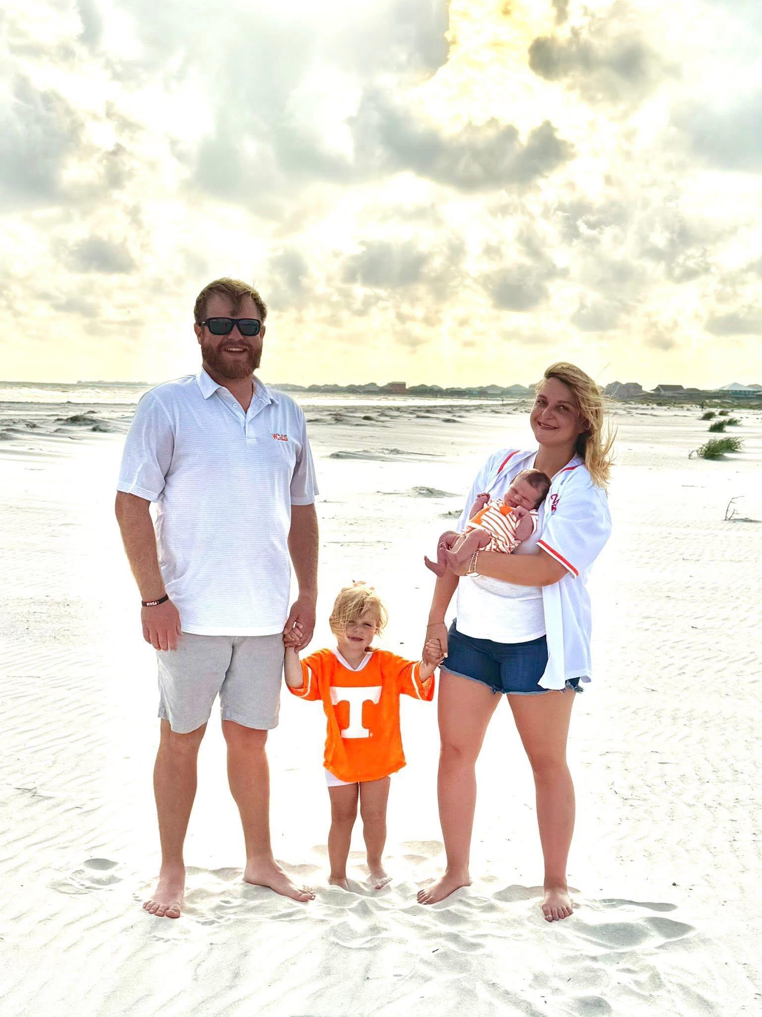 A family of four stands on a sandy beach under a cloudy sky.