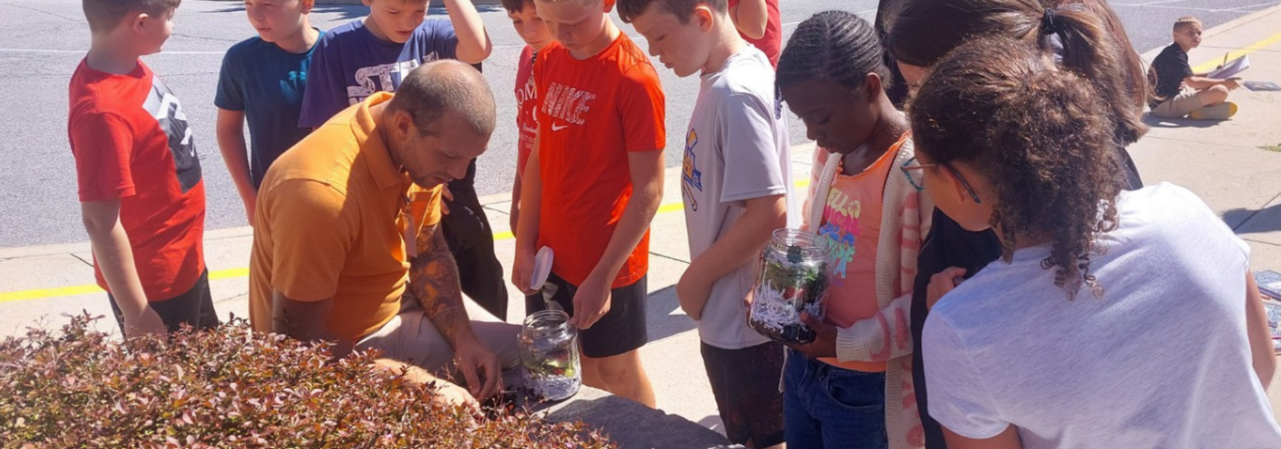 A group of children watching a man while he demonstrates planting in jars.