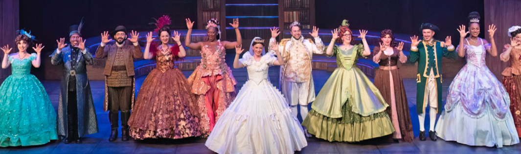 The cast of ZACH Theatre's Cinderella production takes a bow on stage, joined by members of the Austin Deaf Community. The performers wear elegant, colorful costumes, including vibrant ball gowns and regal attire, set against a backdrop of bookshelves and stairs. The cast raises their hands in a gesture of applause, creating an inclusive and celebratory atmosphere.