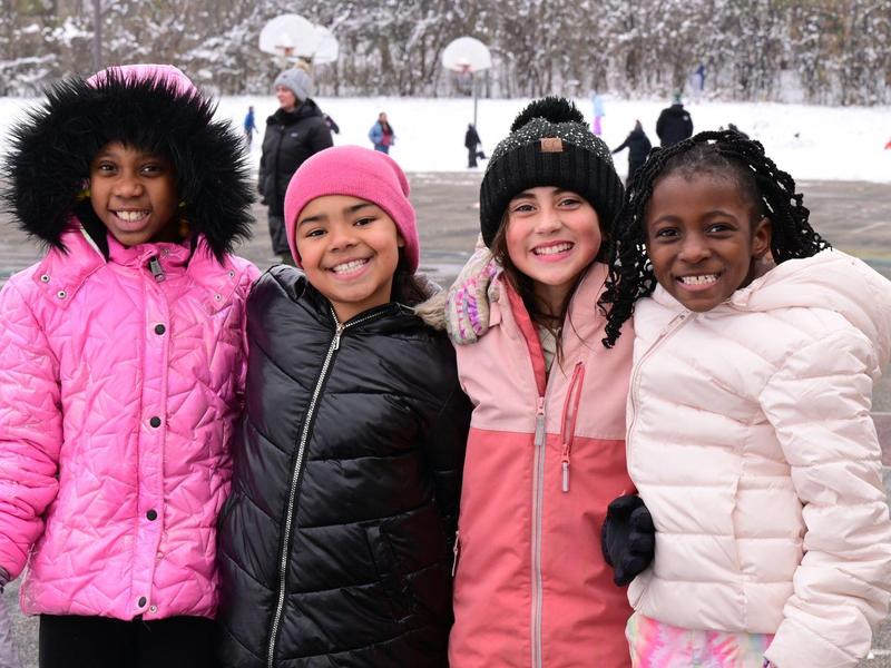 Four children smiling together in winter clothing on a snowy playground.