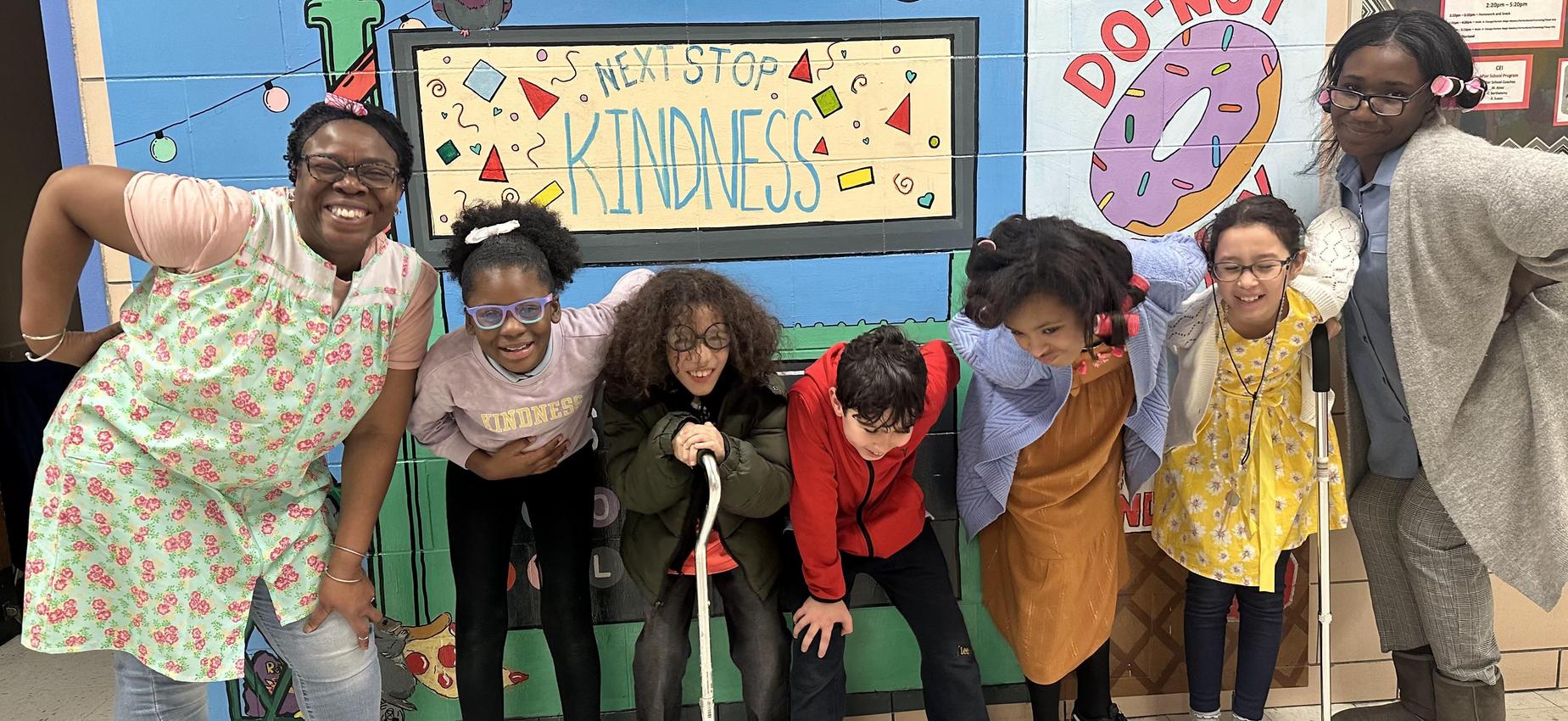 Two female staff members and five students posing in front of the Be Kind school mural on Character Day.