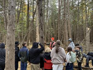 A nature guide speaking to a group of kids in a forest, pointing at a tree.