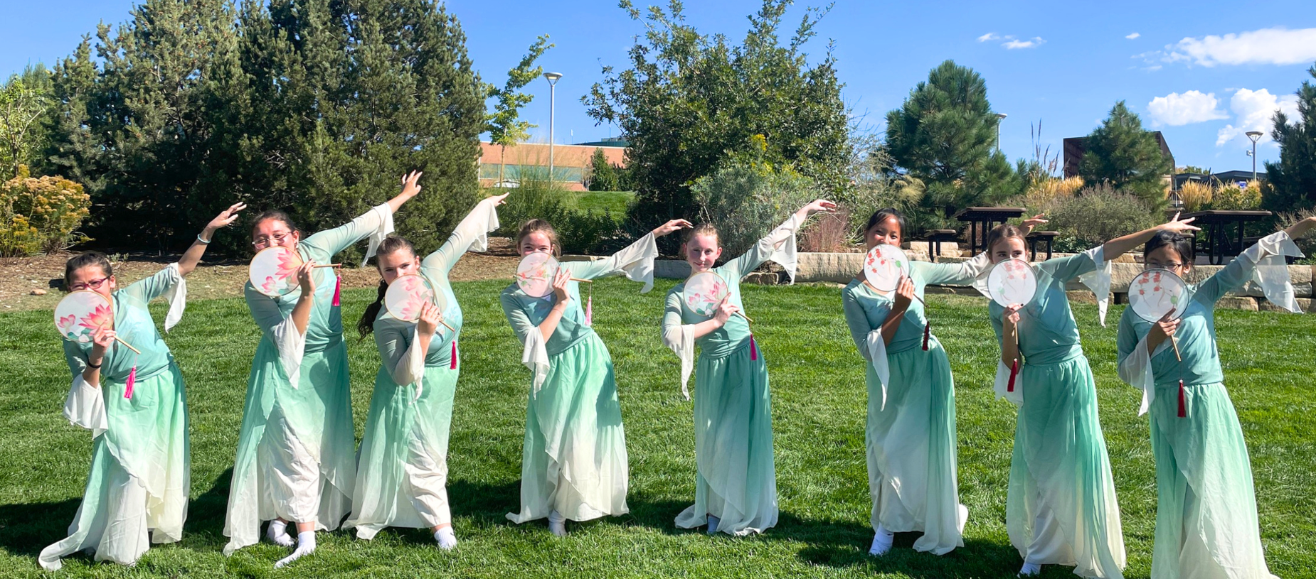 Seven dancers in light green costumes holding fans, posing outdoors on grass.