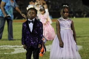 Two young children, one in a purple suit and the other in a dress, stand together.