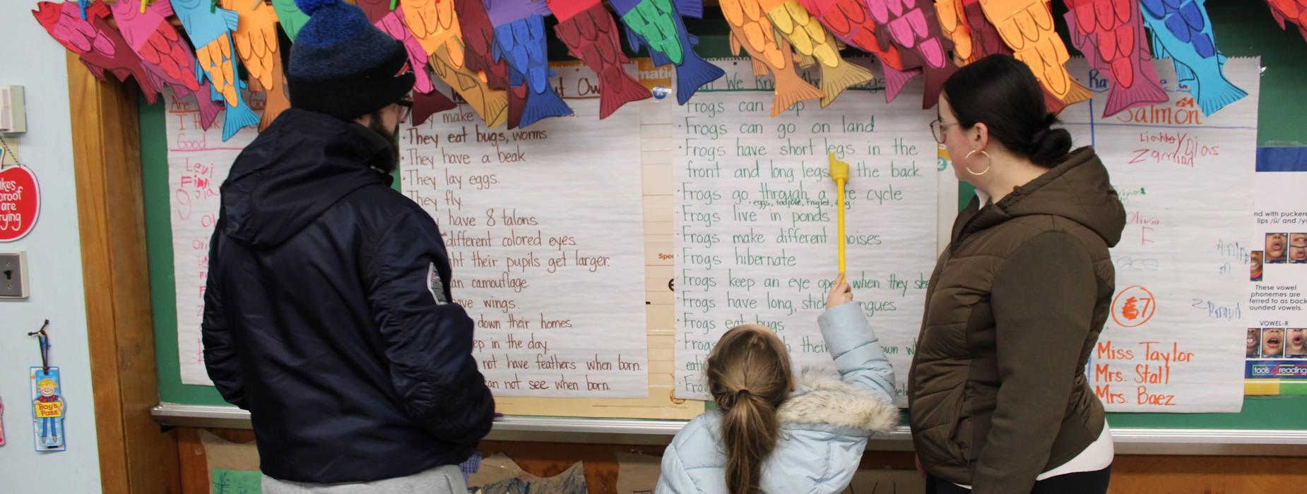 A young student stands holding a yellow pointer, pointing to words on a piece of chart paper. She is reading to two adults, one on either side of her, with a colorful banner of fish above them.