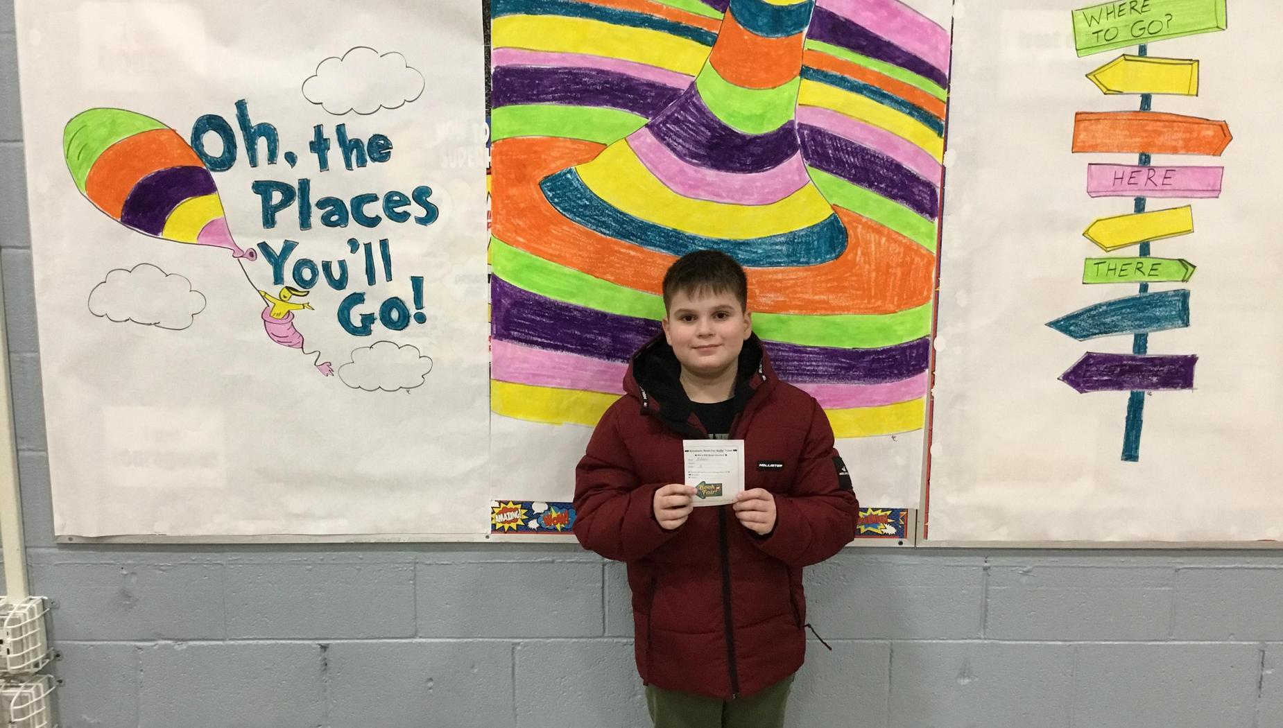 Child in red jacket holding a certificate in front of a colorful mural and signs.