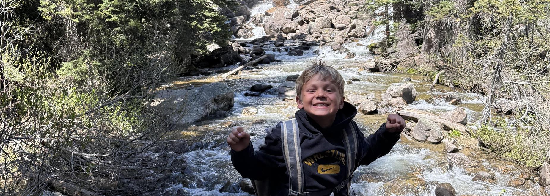 A boy poses joyfully in front of a waterfall.