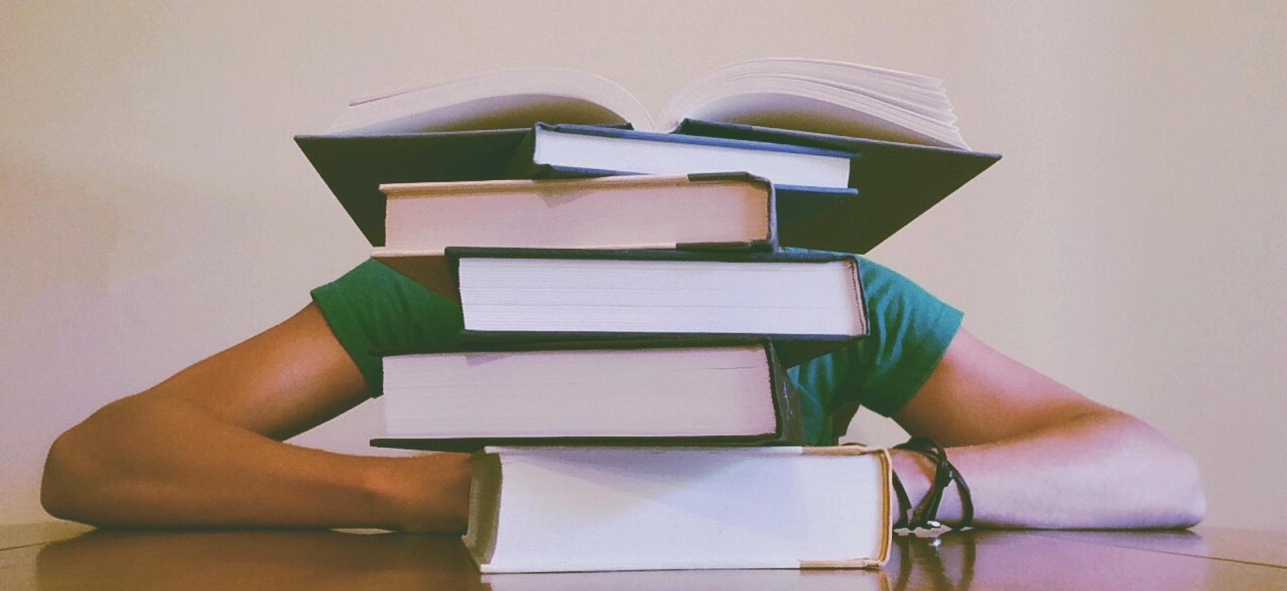 A person sitting at a table with a stack of books obscuring their face.