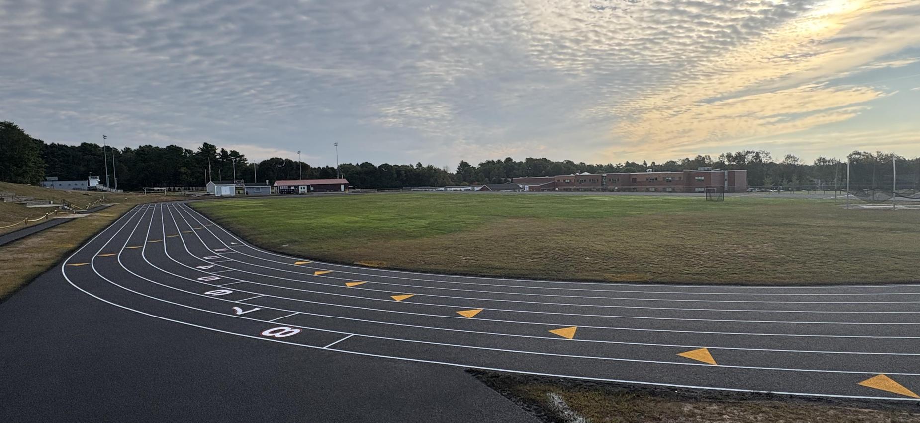 Track and field area with running lanes and a grassy field under a cloudy sky.