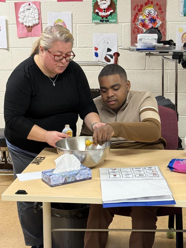 Dr. Kathy helping a student pour food items into a mixing bowl