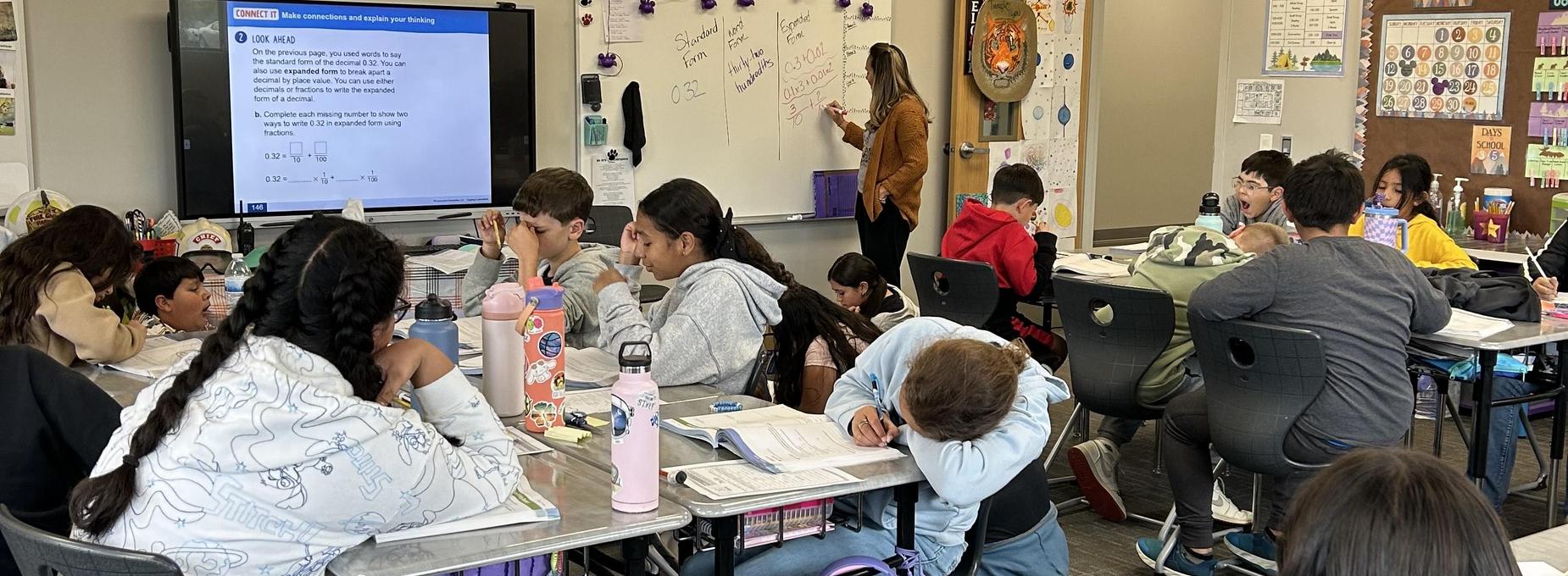 Classroom scene with students working on assignments while a teacher writes on the board.