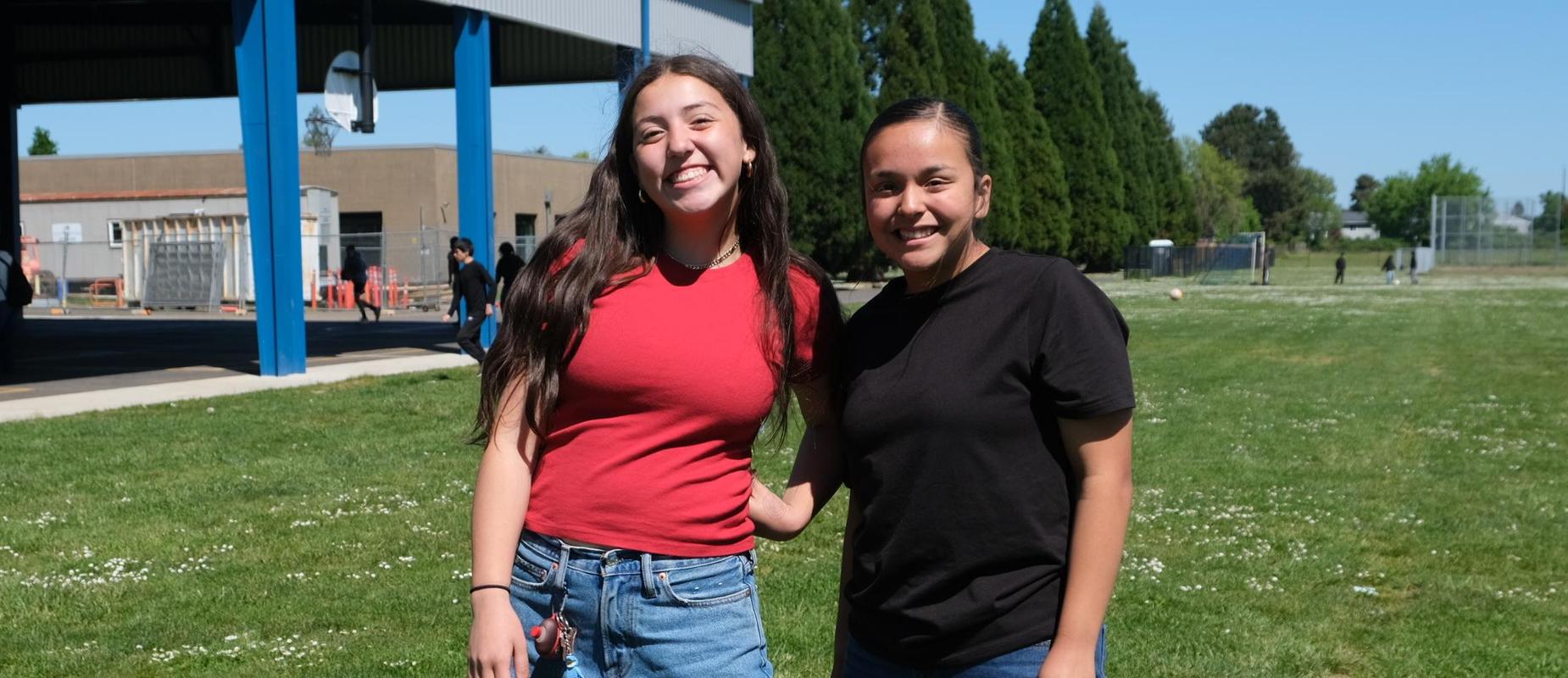 Two girls smiling and posing together on a grassy field in casual outfits.