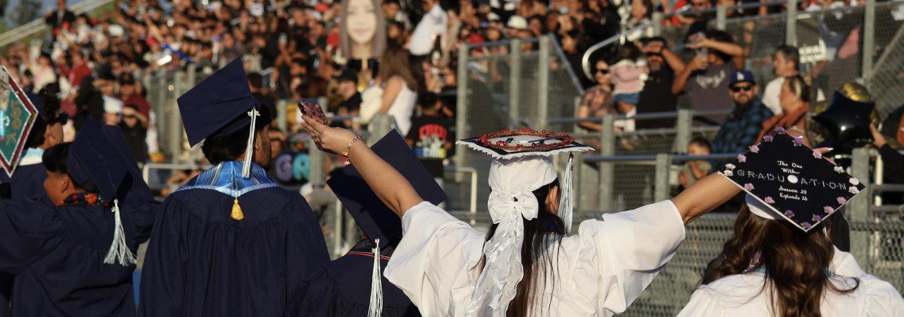 Graduates celebrating at a commencement ceremony, with a cheering crowd in the background.