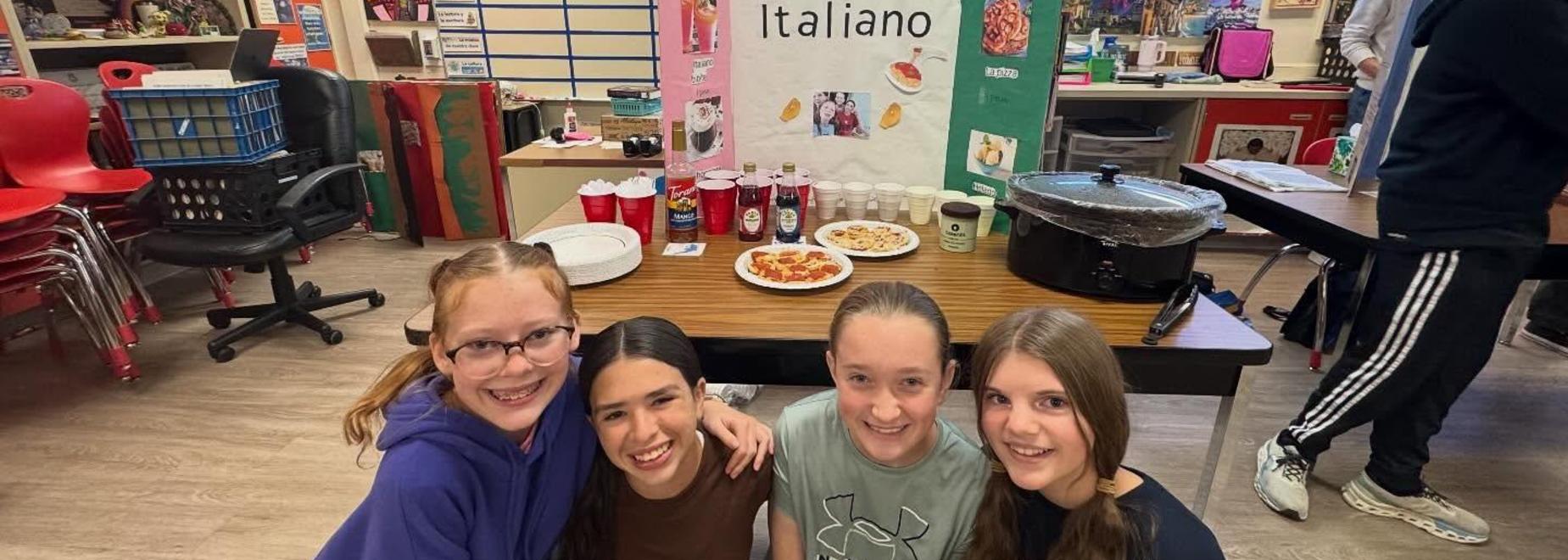 Four girls smile in front of a food display with a poster showcasing Italian cuisine.
