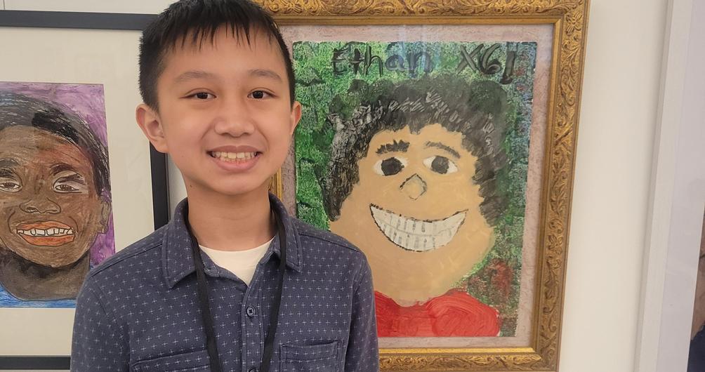 Boy in a blue shirt stands next to a colorful smiling portrait of a boy.