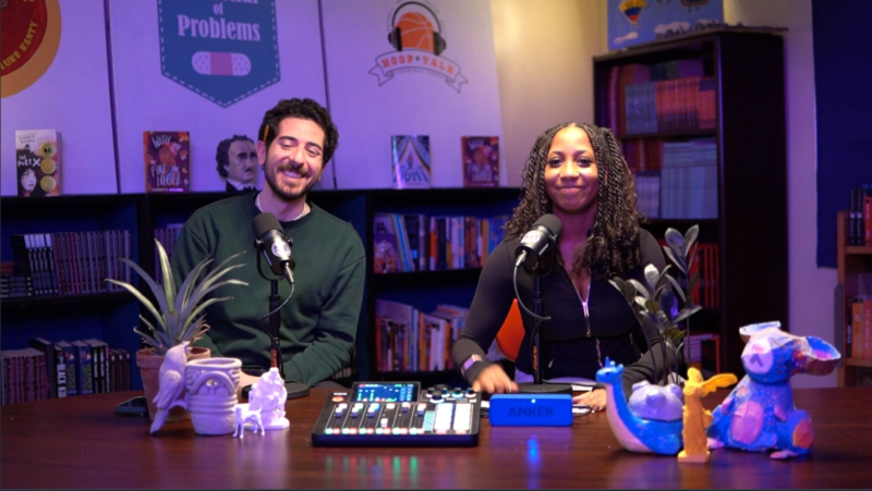 Two people sit at a table with microphones, audio equipment, and art pieces in a podcast studio.