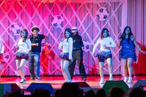 Group of performers dancing on a stage under pink and purple lighting, wearing coordinated outfits with long-sleeve white tops, blue fringe skirts or dresses, and athletic shoes. A few performers in darker clothing stand among them. The backdrop features a lattice pattern decorated with large soccer ball graphics, and audience silhouettes are visible at the bottom of the image.