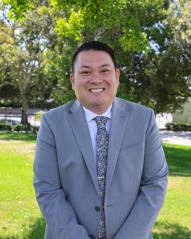 Ryan Shoji standing in a grey suit posing in front of green trees
