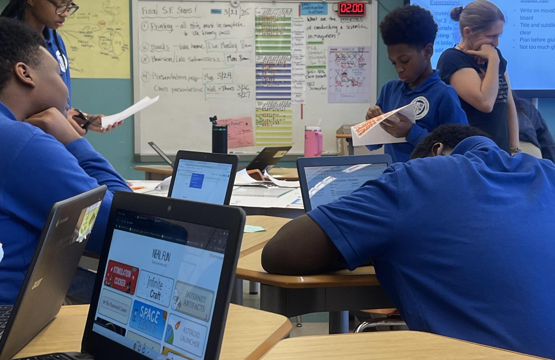 Students engaged in group work with laptops open and a teacher providing instruction.