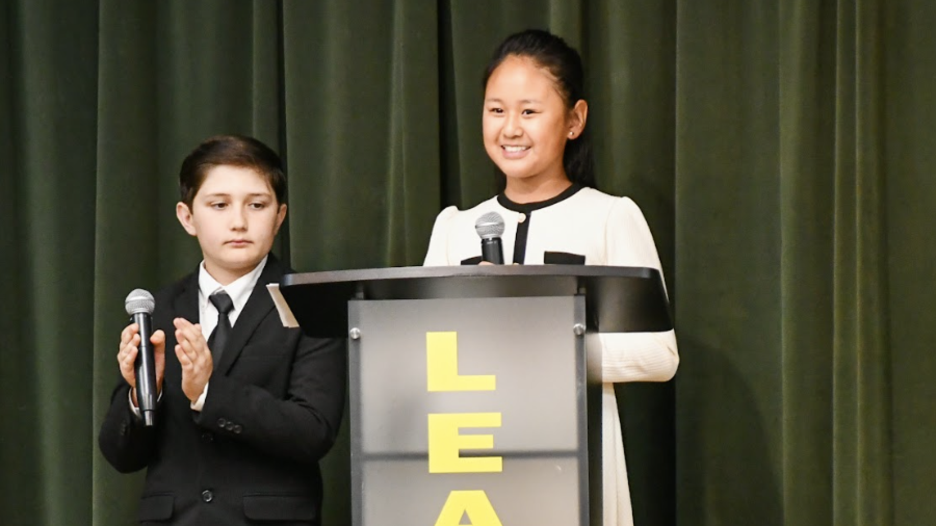 Two individuals stand at a podium labeled 'LEADER' in a formal setting.