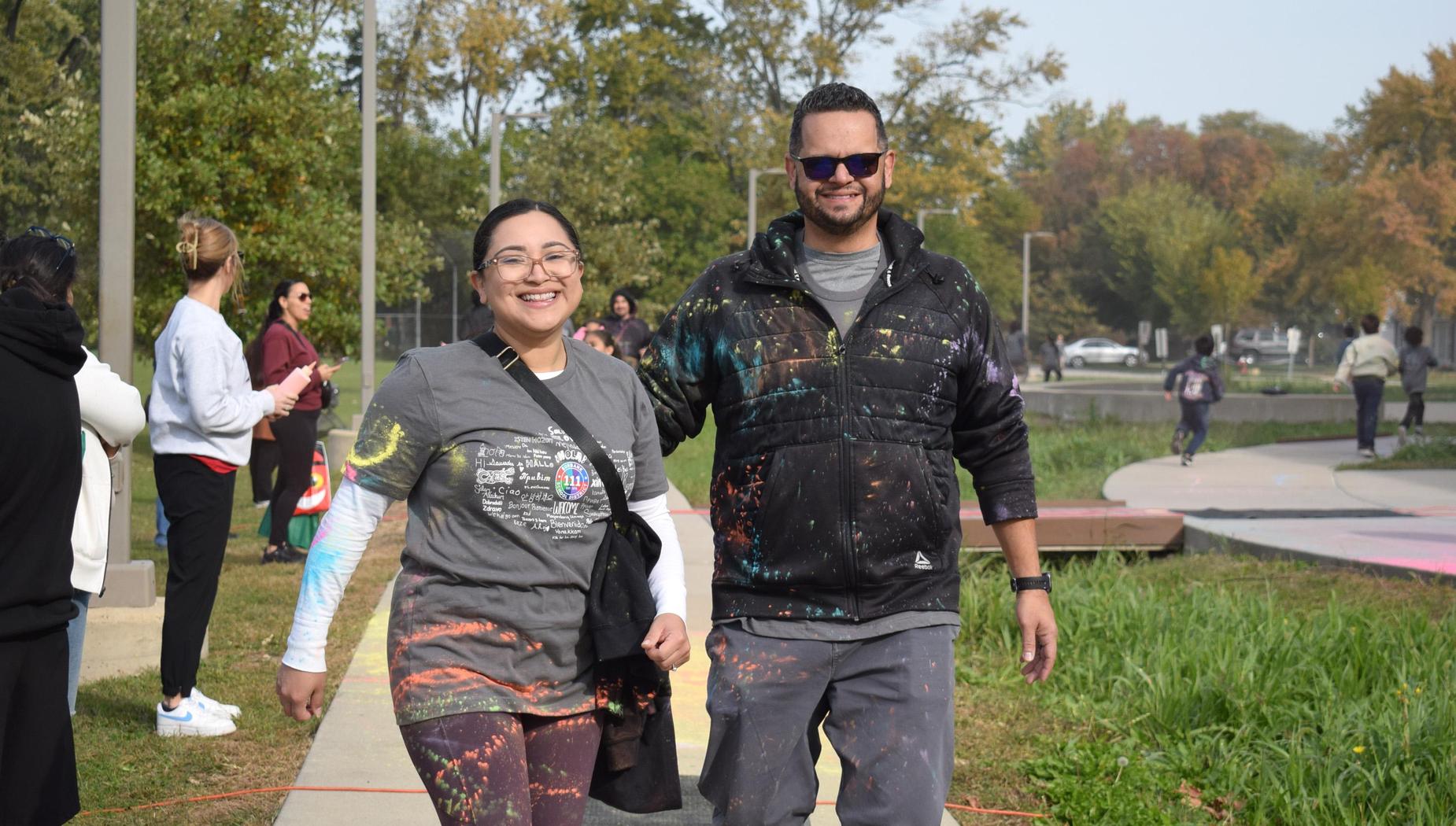 A woman and a man walk together, both covered in vibrant colors.
