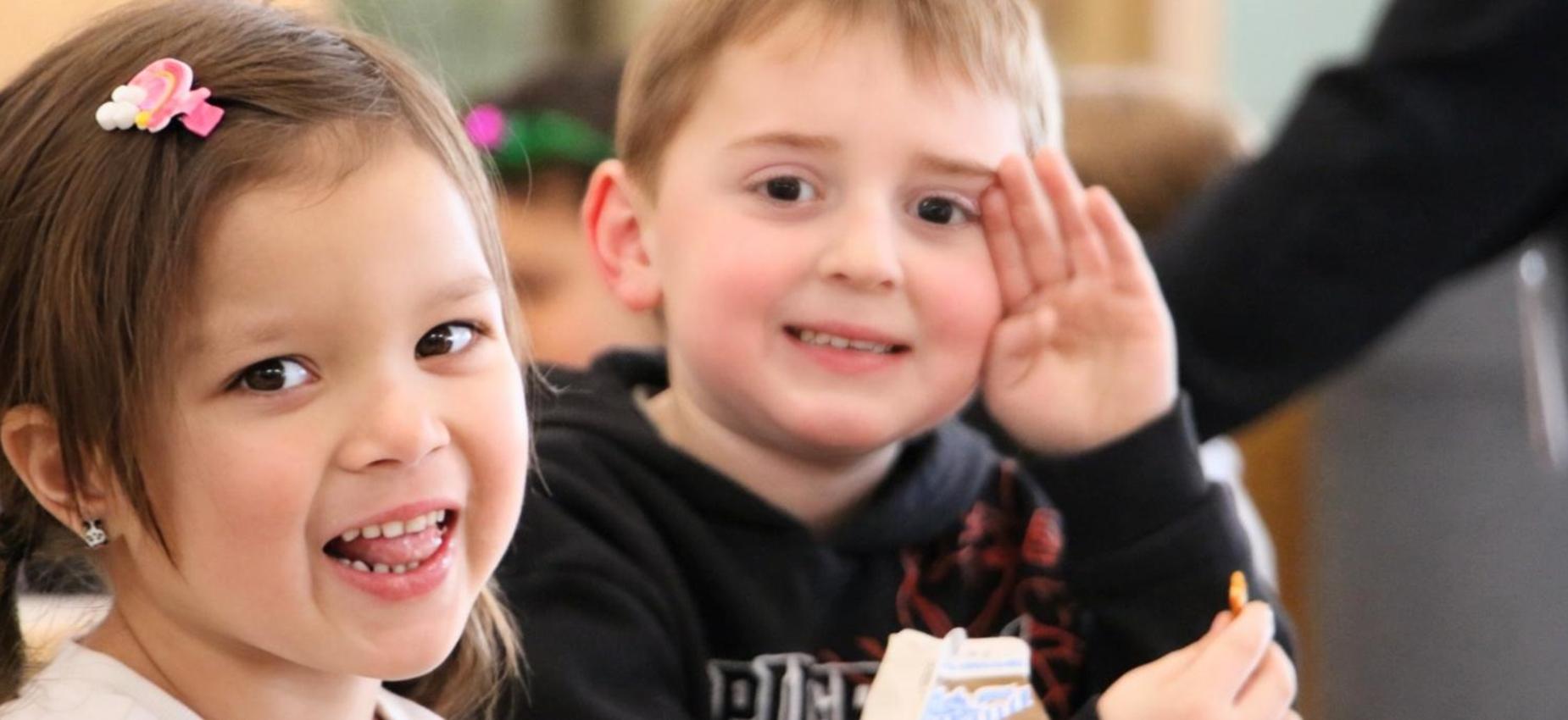 Two children enjoying snacks at a table, one waving and the other smiling.