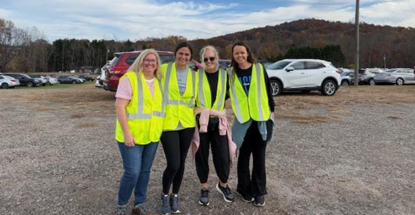 Four women in fluorescent vests smiling outdoors with a green hillside in the background.