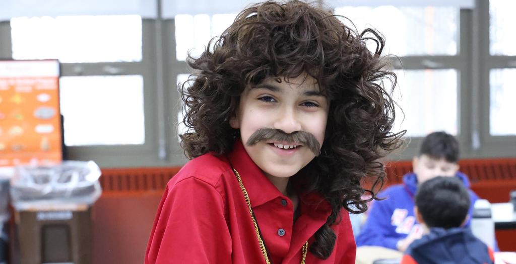 Boy dressed for seventies day, wearing a red shirt, mustache and peace sign emblem around his neck.