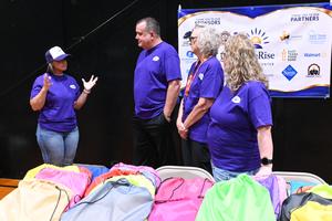 Four adults in purple shirts discussing in front of a sponsor banner and colorful bags.