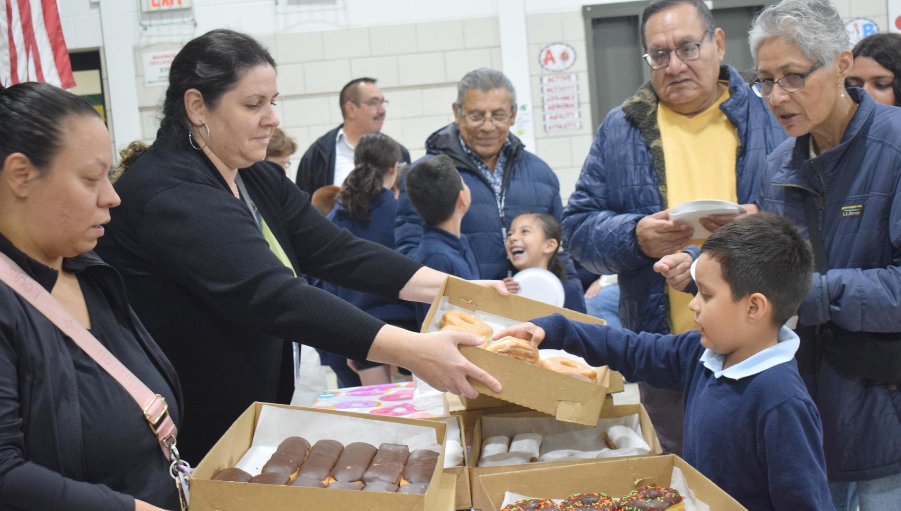 An adult hands a box of donuts to a young boy while others enjoy treats.