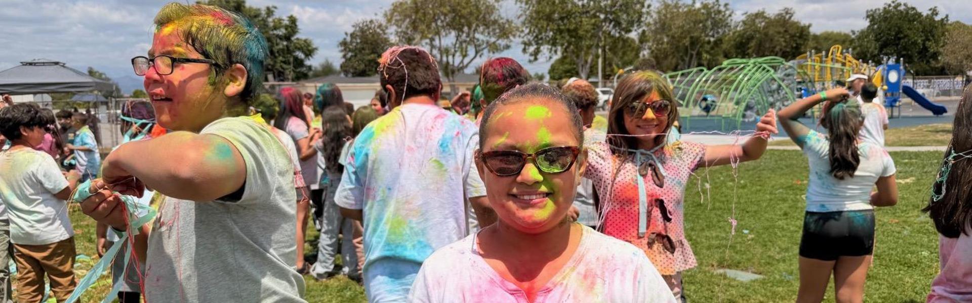 Group of children covered in colorful powders enjoying an outdoor celebration.