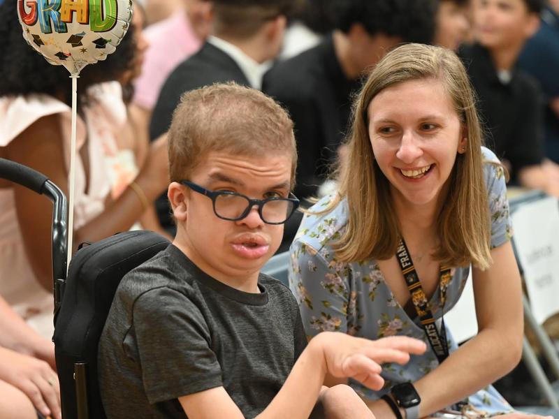 Boy in a wheelchair smiles alongside a woman at a graduation event.