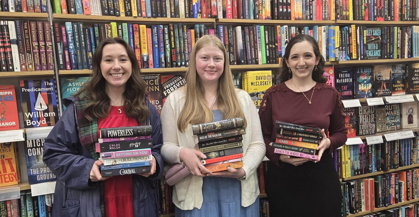Three students holding stacks of books in a l bookstore aisle.