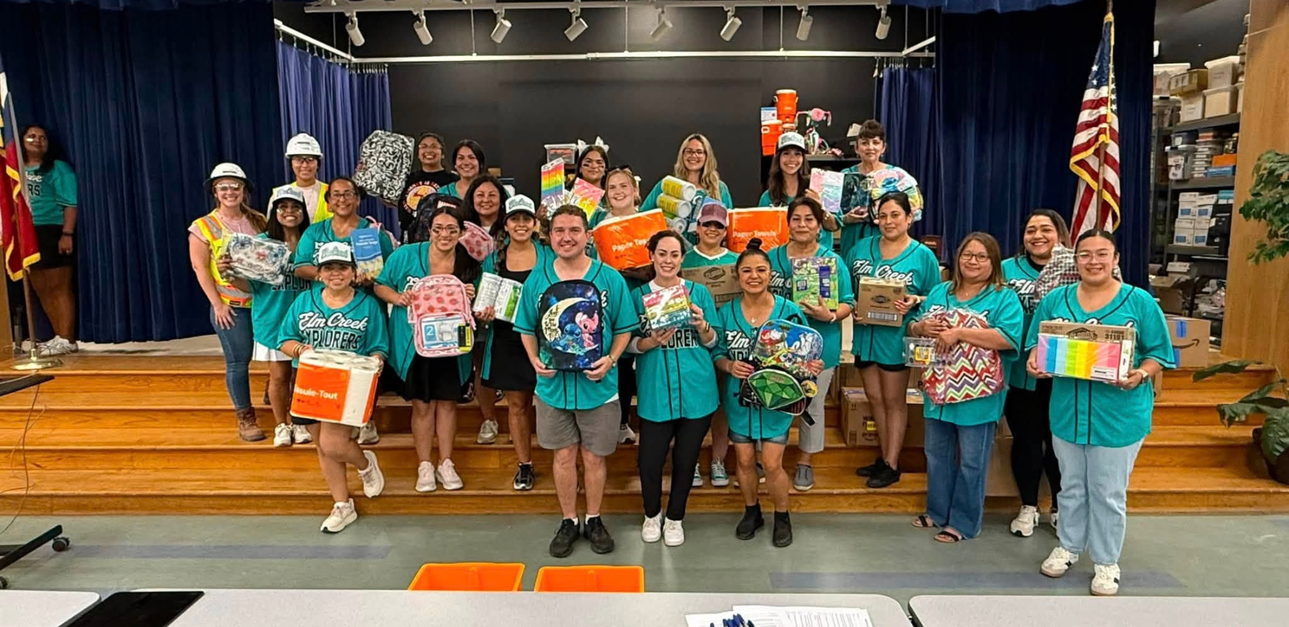 Team of volunteers in matching shirts holding supplies on a stage in a school auditorium.