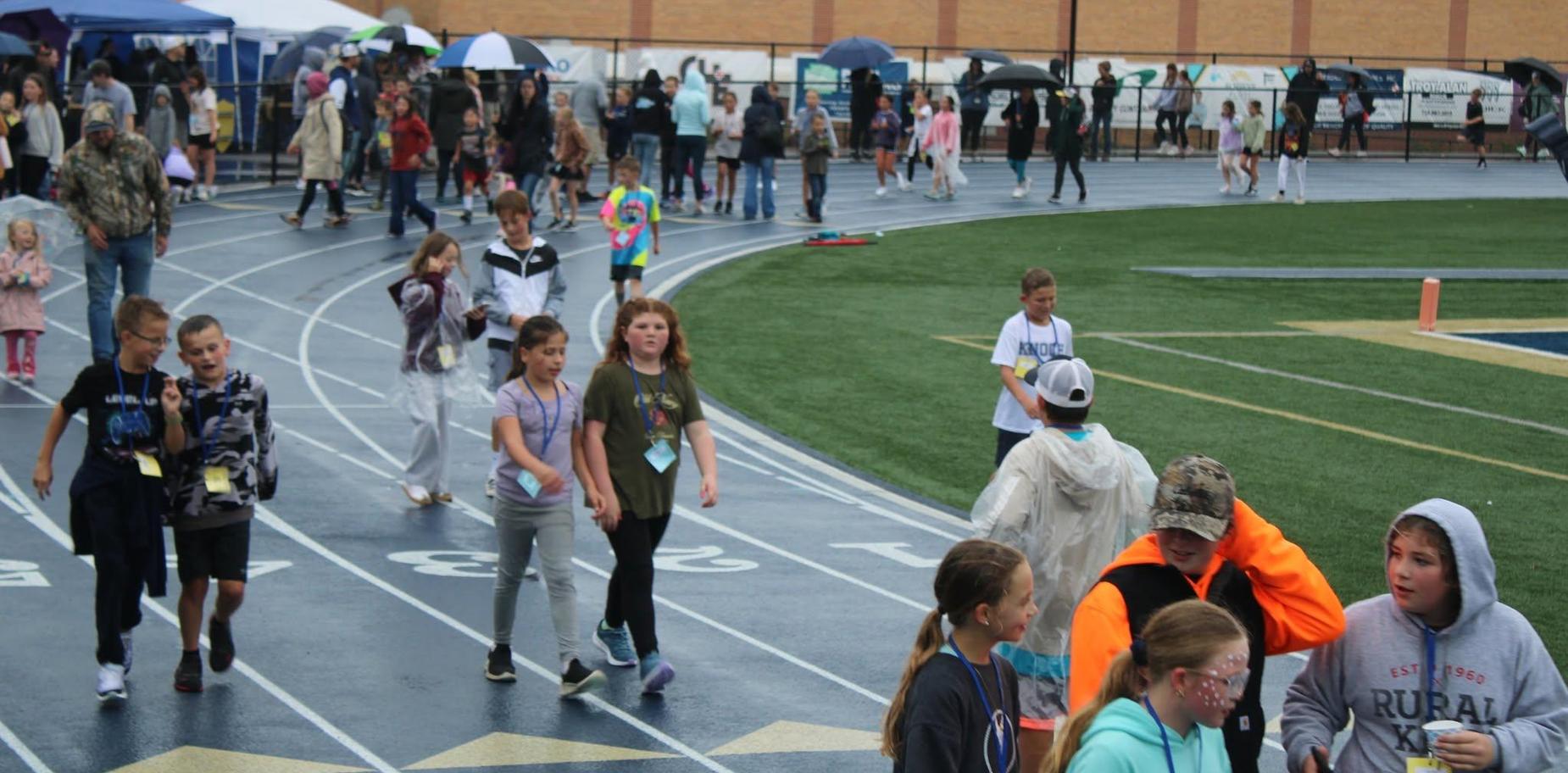 Children walking on a track at an outdoor event, some with umbrellas in the rain.