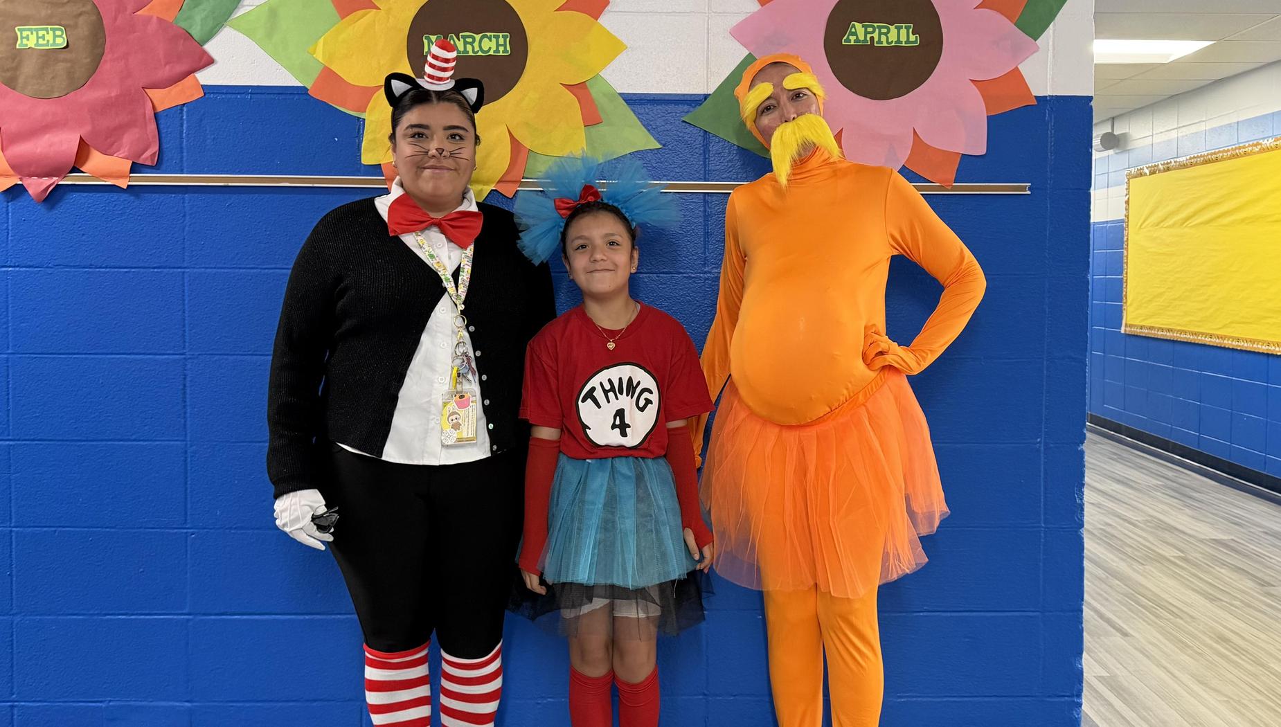 Three individuals in costumes pose in front of flower decorations on a blue wall.