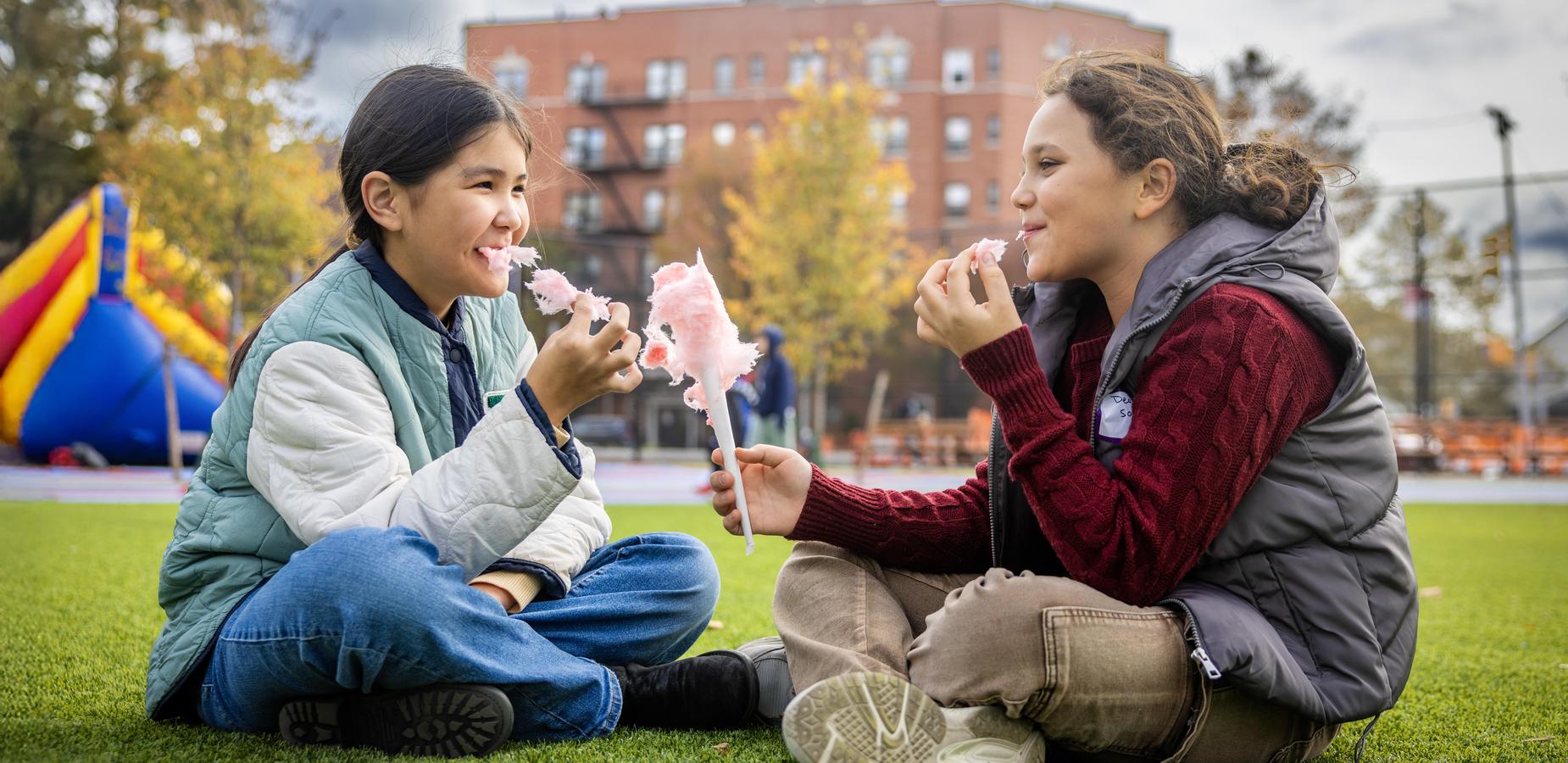 Two girls sitting on grass enjoying cotton candy while smiling.