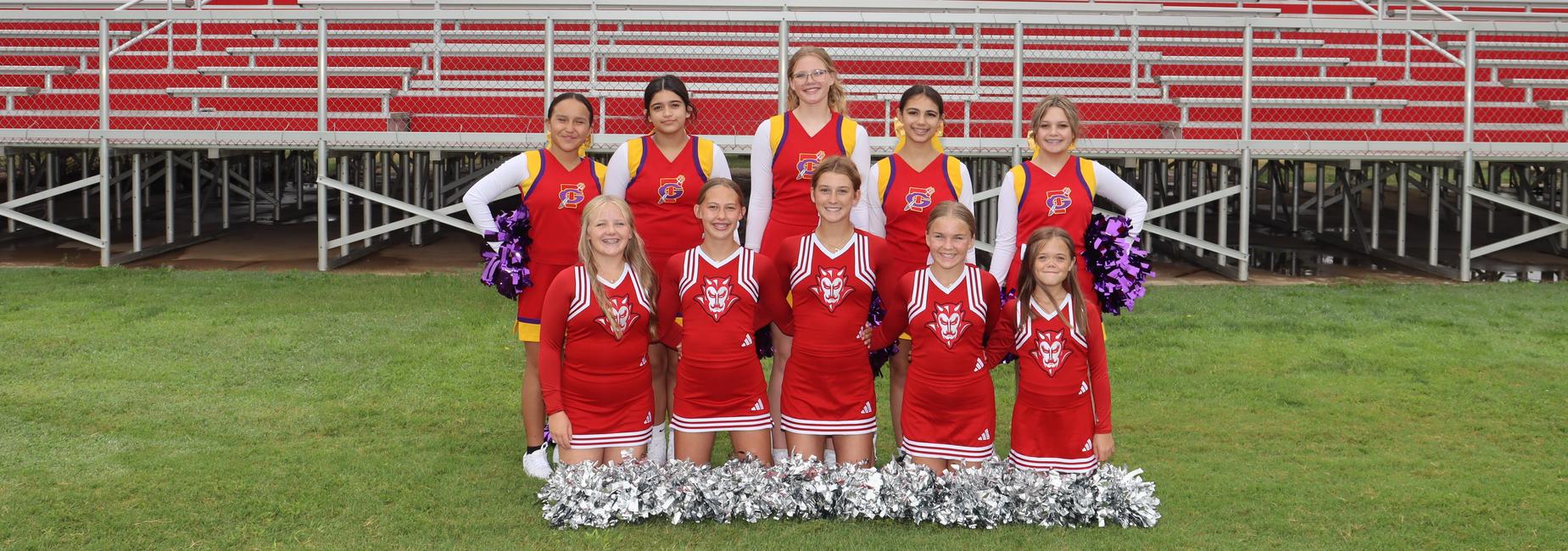 A group of young cheerleaders in red uniforms with purple pom-poms, posed in front of bleachers.