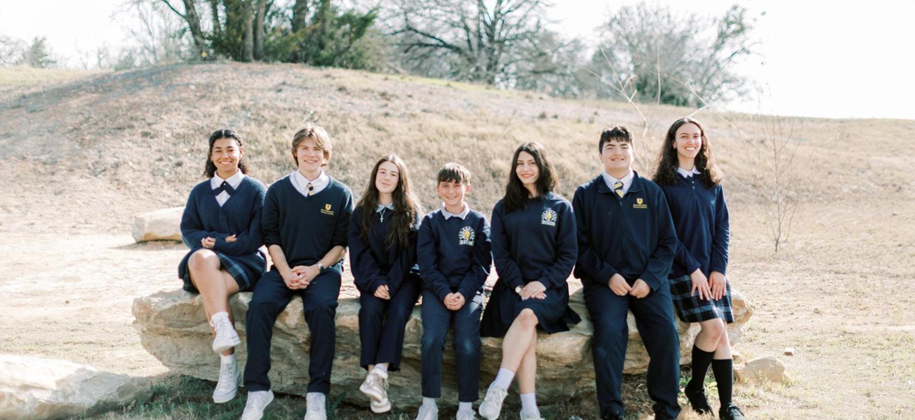 Seven upper school students in uniform sit together on a rock outside