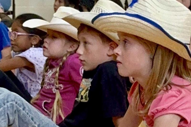 A group of children in cowboy hats sit on the gym floor, watching the assembly with focused expressions.