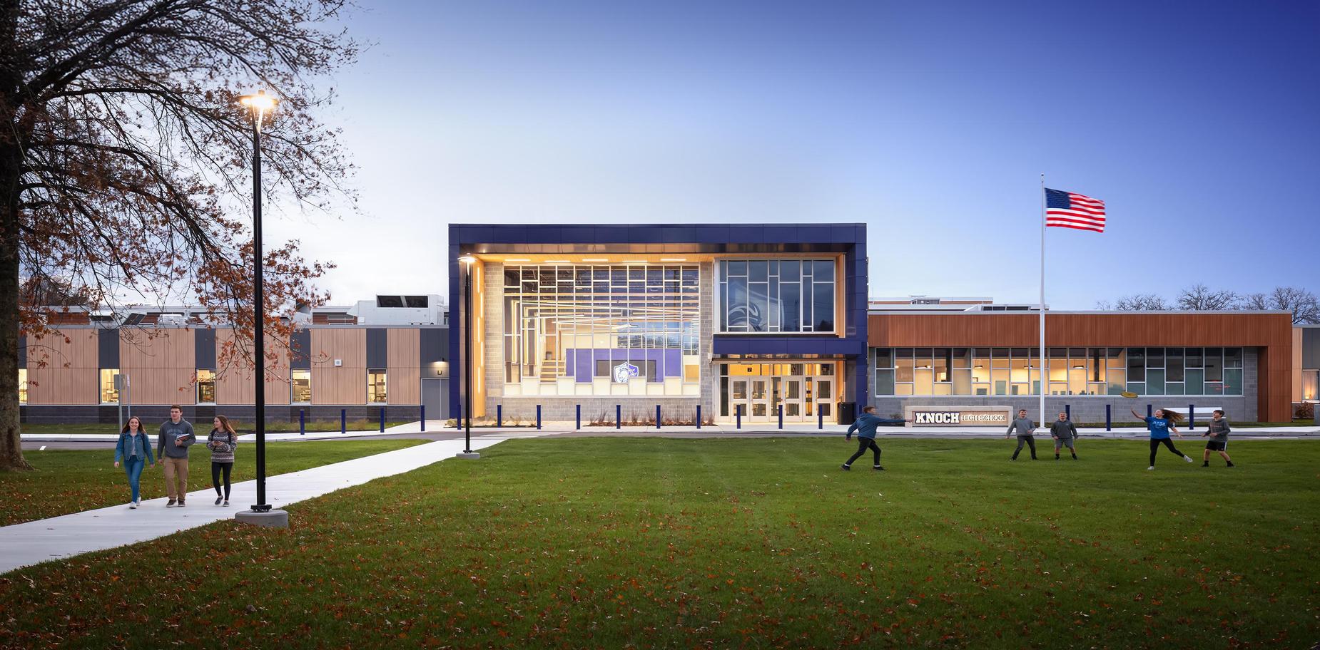 brightly lit high school with flag flying beside the school and students walking on campus
