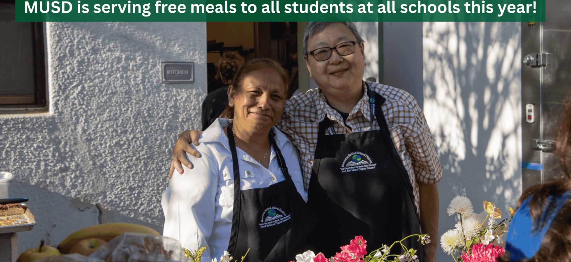 Two smiling women in aprons at a table with food, promoting free meals for students.